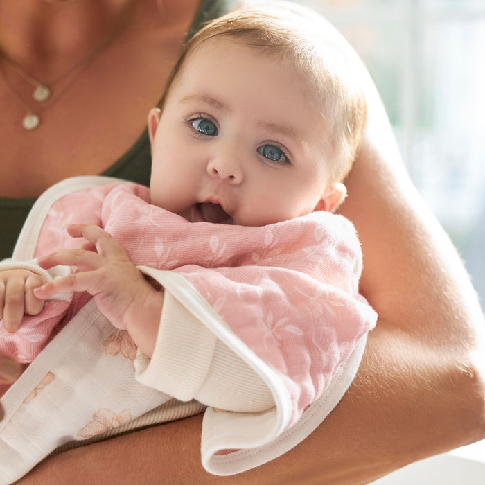 A baby with big blue eyes is wrapped in a pink and white towel, held in loving arms. The softly blurred background highlights the cozy vibe of HALOs Mini Crib Essentials Bundle—perfect for soothing newborn bedtime moments.