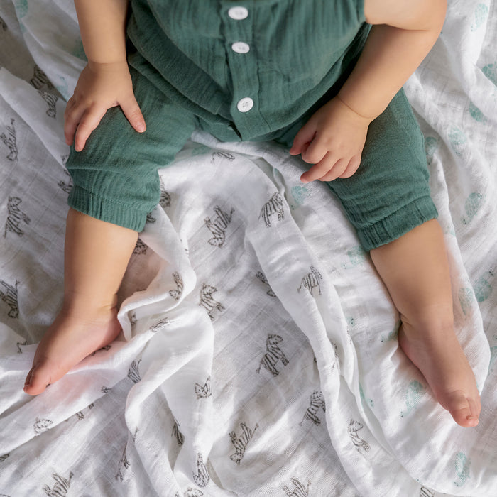 A baby sits on a white blanket featuring animal prints, wearing a green outfit. The blanket is the aden + anais organic cotton muslin burpy bibs 2 pack. The photo is taken from above, showing the baby's legs stretched out and one hand resting on their knee.