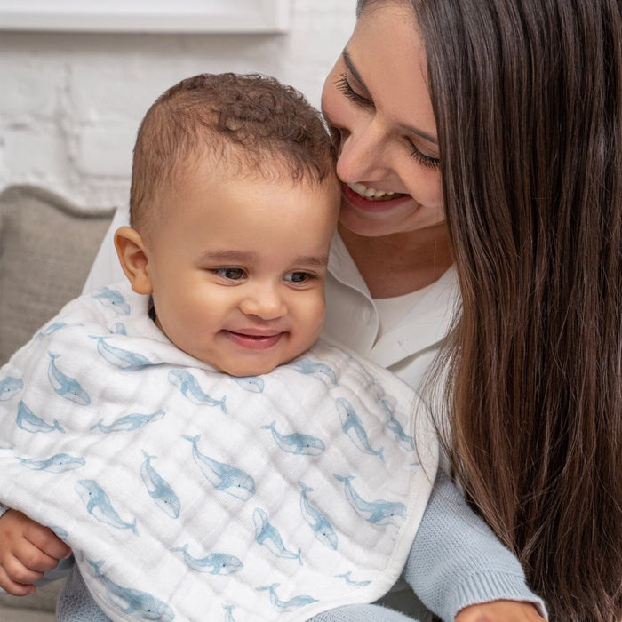 A woman smiles at a cheerful baby wrapped in a whale-patterned blanket. They share a joyful moment on the couch, with aden + anais organic cotton muslin burpy bibs nearby, adding gentle comfort.