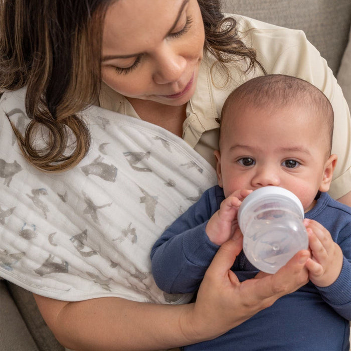 A woman cradles her baby tenderly on the couch as the little one drinks from a bottle. Draped over her shoulder is HALOs Welcome Home Bundle, an essential for newborns. Her gaze radiates affection and warmth.