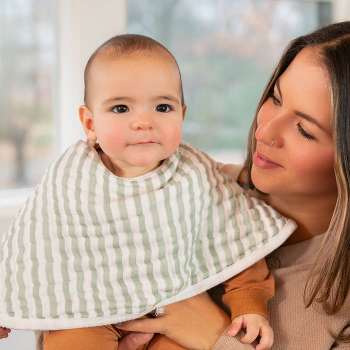 A woman with long hair lovingly holds a baby who is wearing a striped bib. The baby, resting on aden + anais organic cotton muslin burpy bibs, gazes neutrally at the camera in a softly lit indoor setting with a blurred background.