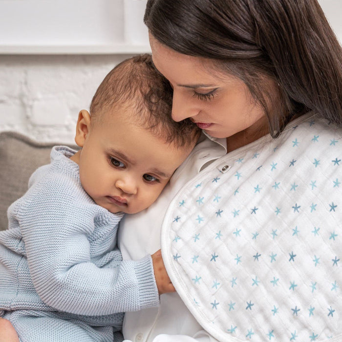 A woman, wearing a white top, holds a baby in a light blue knitted outfit on the couch. The baby leans on her shoulder while she uses an aden + anais organic cotton muslin burpy bib featuring blue stars. Both seem calm and content.