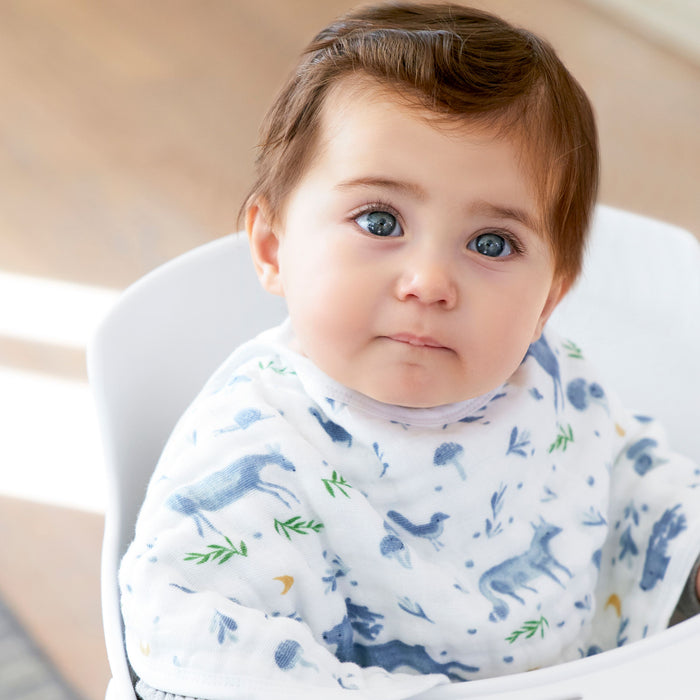A baby with brown hair and blue eyes sits in a high chair, wearing an aden + anais organic cotton muslin burpy bib featuring blue animal prints. The GOTS-certified fabric enhances the serene atmosphere as the baby looks up peacefully in the softly lit room.