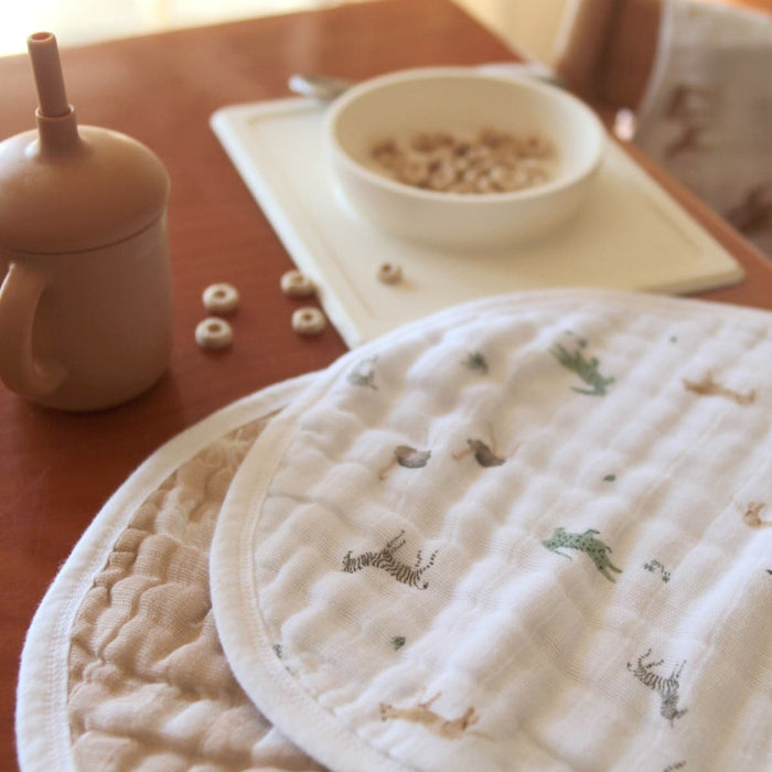 A close-up of a wooden table showcases aden + anais organic cotton muslin burpy bibs, GOTS certified, alongside a round sipping cup with a straw and a bowl of cereal on a white placemat. Scattered cereal pieces add charm to the breakfast scene.