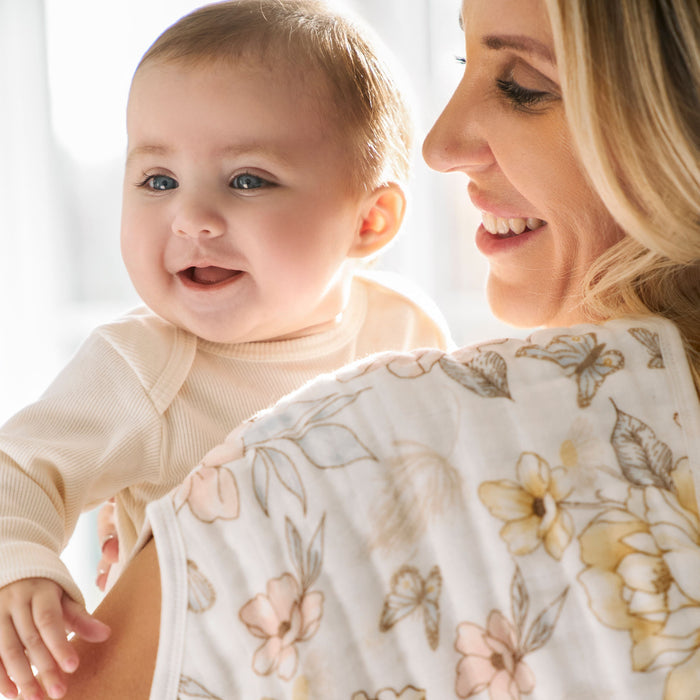 A blonde-haired woman smiles while holding a laughing baby snugly wrapped in an aden + anais organic cotton muslin burpy bib. The baby, in a light outfit, leans over her shoulder as she wears a floral cloth, with the background softly blurred.