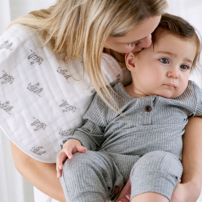 A woman gently kisses a baby in a gray outfit who looks calmly into the distance, with a zebra-patterned aden + anais organic cotton muslin burpy bib draped over her shoulder.