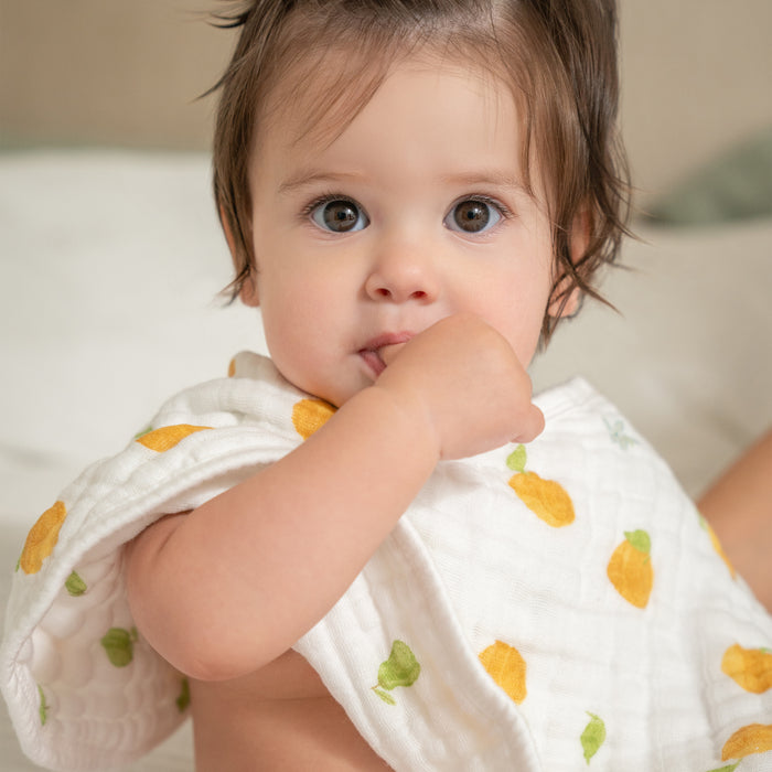 A baby with big eyes and dark hair sits with a hand in their mouth under a white blanket with yellow and green patterns. They are nestled on the HALOs Smart BassiNest Bundle, enjoying comfort against a softly blurred background.