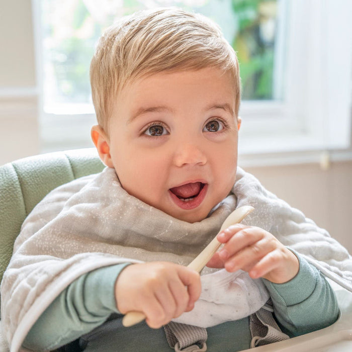A happy baby sits in a high chair, wrapped in a patterned blanket, holding a spoon with both hands. The blonde child wears a green shirt as bright windows reveal lush greenery outside—a serene scene perfect for the HALO Smart BassiNest Bundles AutoSoothe™ feature to lull the little one to sleep.