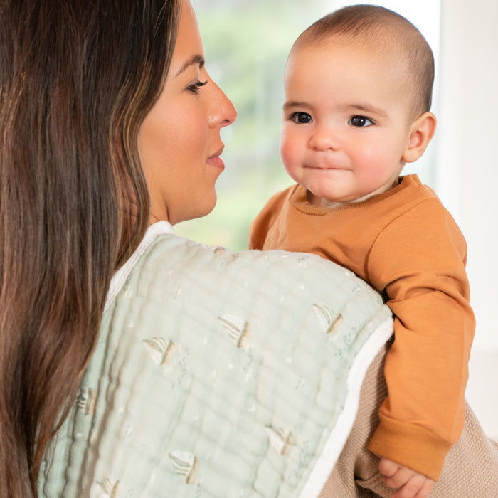 A woman with long brown hair holds a smiling baby in a patterned cloth, wearing a brown shirt. She wears a light-colored top and drapes an aden + anais organic cotton muslin burpy bib from the 2 pack over her shoulder. The background is softly blurred.