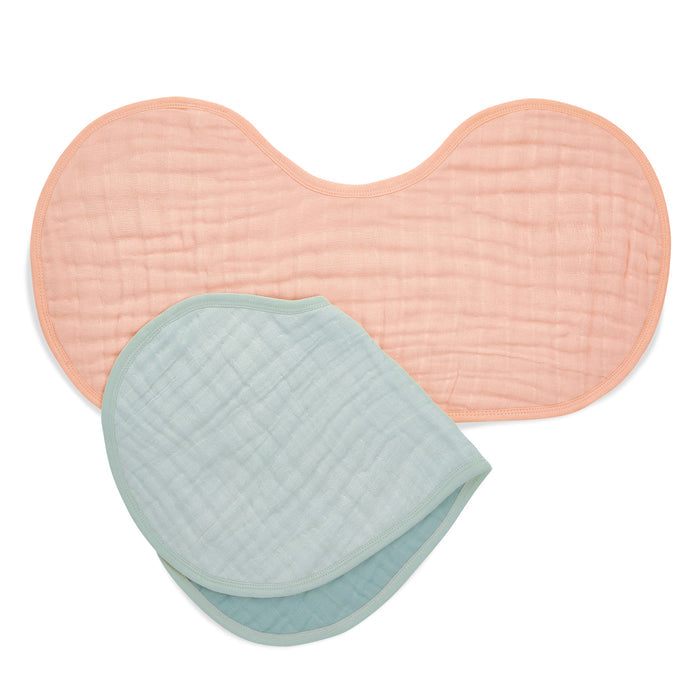 Two aden + anais organic cotton muslin burpy bibs are displayed. The top bib is light pink, hourglass-shaped, while the bottom one is folded and light green, both featuring a soft texture and GOTS certification for comfort.