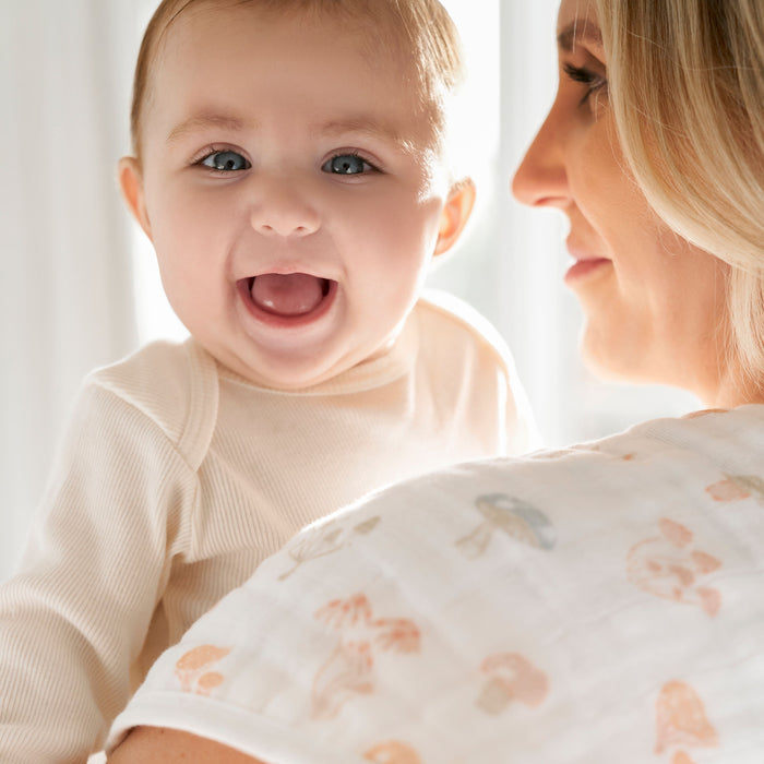 A smiling baby in a cream outfit is held by a blonde woman with an aden + anais white patterned blanket over her shoulder. A nearby organic cotton muslin burpy bib from the aden + anais 2 pack completes the warmly lit, cheerful scene.