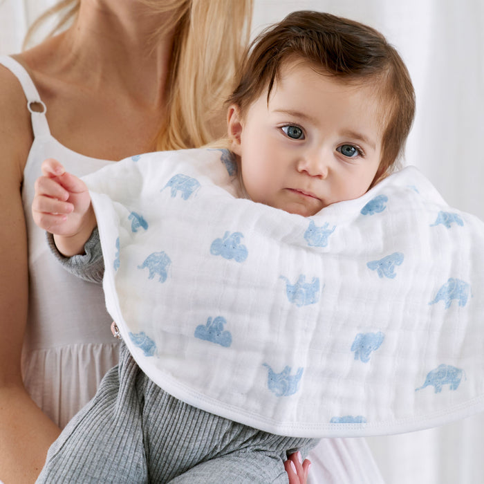 A baby with light brown hair is held by an adult in a white sleeveless top. The baby wears a gray outfit and a white bib with blue elephants, matched with aden + anais GOTS certified organic cotton muslin burpy bibs 2 pack, as the soft-focus background enhances the gentle scene.