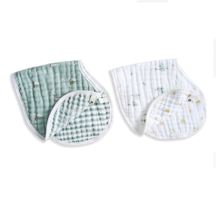 Two aden + anais organic cotton muslin burpy bibs are shown: one in light teal with small prints and a checkered pattern on the reverse; the other is white with tiny floral designs and light teal patterns on the reverse. Both feature quilted textures, snap closures, and GOTS certified cotton.