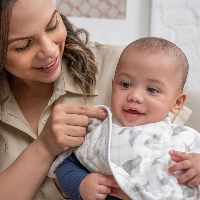A smiling adult lovingly gazes at a happy baby wrapped in a patterned blanket. The baby looks forward while indoors, surrounded by warmth. Nearby are aden + anais organic cotton muslin burpy bibs, adding to the cozy ambiance.