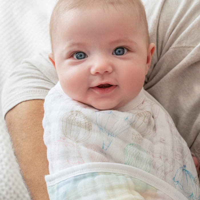 A smiling baby with blue eyes is wrapped in an aden + anais patterned organic cotton muslin blanket, held by an adult. The background is softly blurred.