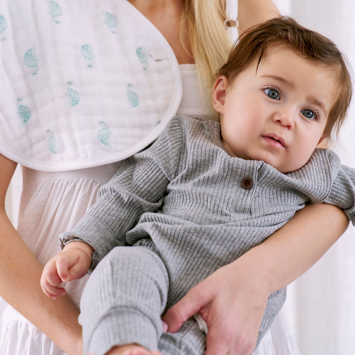 A person in a white dress gently cradles a baby in a gray outfit. An aden + anais organic cotton muslin burpy bib with blue patterns rests on their shoulder, enhancing the soft, serene atmosphere.