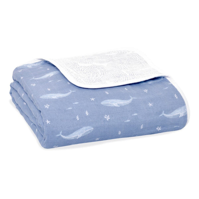 The aden + anais organic cotton dream blanket is a folded blue blanket with white whale patterns and a soft white edge, made from GOTS certified cotton muslin. It features a textured white interior visible at the corner.