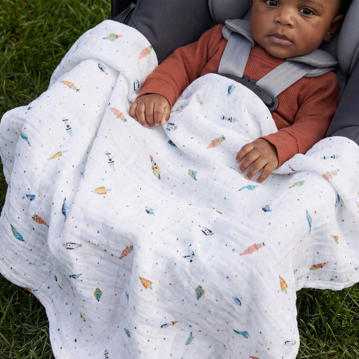A baby in a brown outfit sits in a car seat on grass, covered with an aden + anais organic cotton muslin dream blanket featuring small, colorful patterns.
