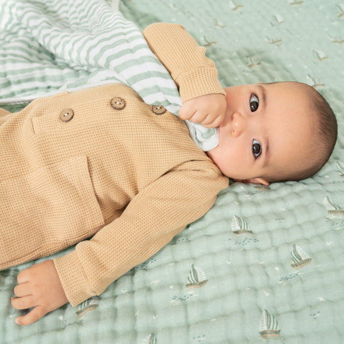 A baby in a tan outfit is lying on an aden + anais organic cotton muslin dream blanket with small sailboat patterns. The textured green backdrop is soft, GOTS certified cotton muslin. The baby holds a green and white striped cloth, gazing upward with curiosity.