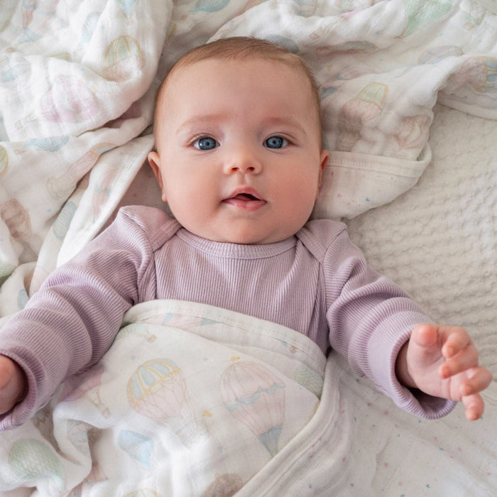 A blue-eyed baby in lavender attire lies on a bed partly covered by the aden + anais organic cotton dream blanket, featuring balloon patterns. The cozy muslin fabric envelops the pastel bedding as the baby gazes upward with hands slightly raised.