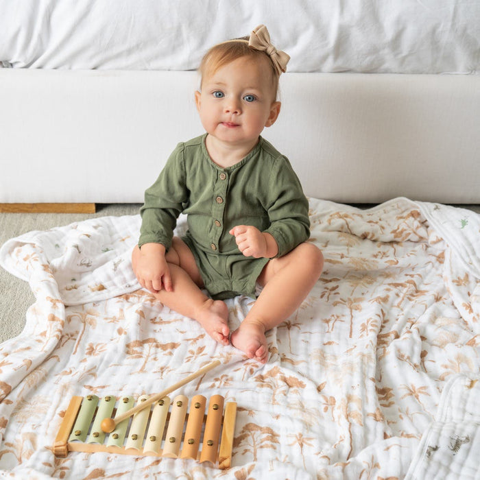 A baby with a bow plays adorably on an aden + anais organic cotton dream blanket, wearing a green outfit. The soft, floral-patterned muslin offers comfort as the baby enjoys a colorful wooden xylophone and gazes at the camera.