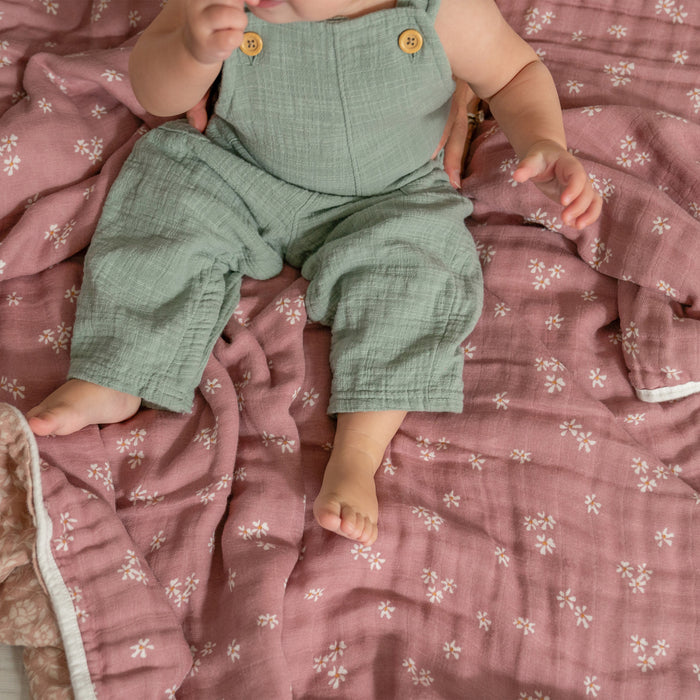 A baby in a green romper sits on an aden + anais organic cotton muslin dream blanket featuring small white floral patterns. The GOTS certified pink blanket is crumpled cozily, with the babys hand raised and feet visible.