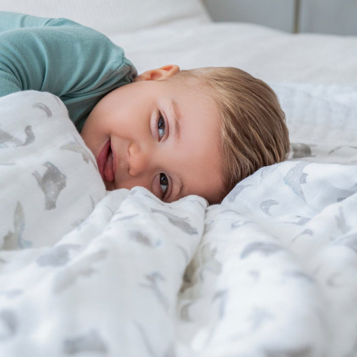 A smiling blond toddler lies on a bed, partially covered by an aden + anais organic cotton dream blanket with gray animal prints. Wearing a teal shirt and looking toward the camera, the child creates a playful and cozy atmosphere.
