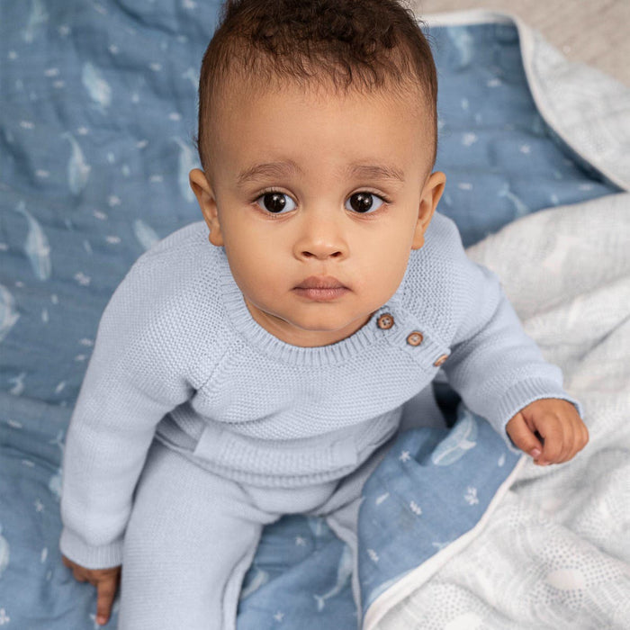 A baby in a light blue knitted outfit sits on an aden + anais organic cotton dream blanket, GOTS certified and adorned with a fish pattern, looking up curiously. The scene is cozy and softly lit.