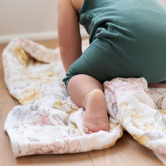 A baby in a green outfit crawls on a wooden floor, partially on an aden + anais organic cotton dream blanket adorned with pastel flowers. The focus is on the babys softly lit legs and feet as they explore the comforting texture of GOTS-certified cotton muslin.