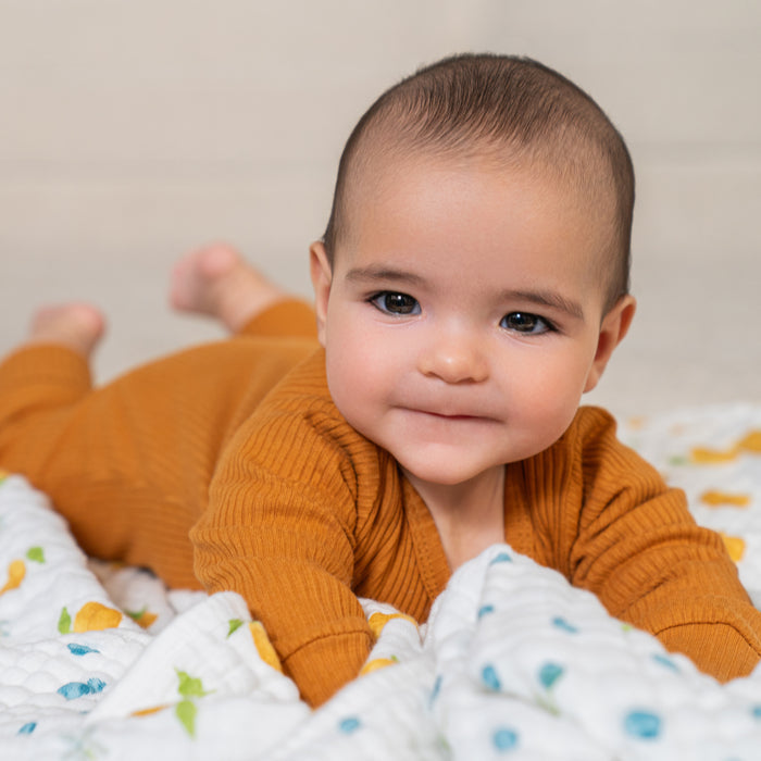A baby in a rust-colored outfit smiles gently while lying on an aden + anais organic cotton muslin dream blanket, propped up on their arms.