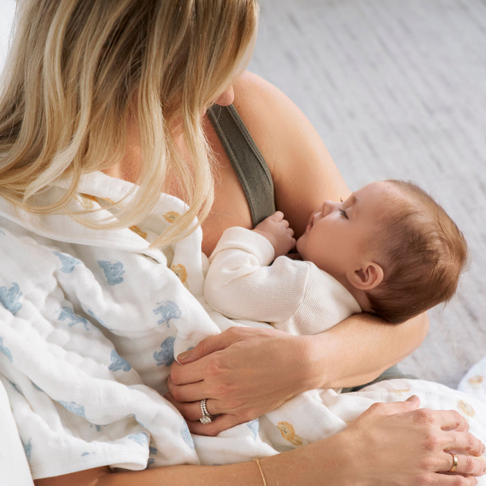 A woman is breastfeeding a baby wrapped in an aden + anais organic cotton dream blanket with blue and yellow animal patterns. They sit on a light-colored surface in a softly lit room, ensuring comfort and warmth. The womans long blonde hair obscures her face.