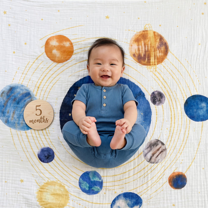 A smiling baby in a blue outfit sits on the aden + anais organic cotton muslin dream blanket, featuring colorful planets and gold orbits. A wooden disc reading "5 months" adds charm to this cozy crib bedding scene.