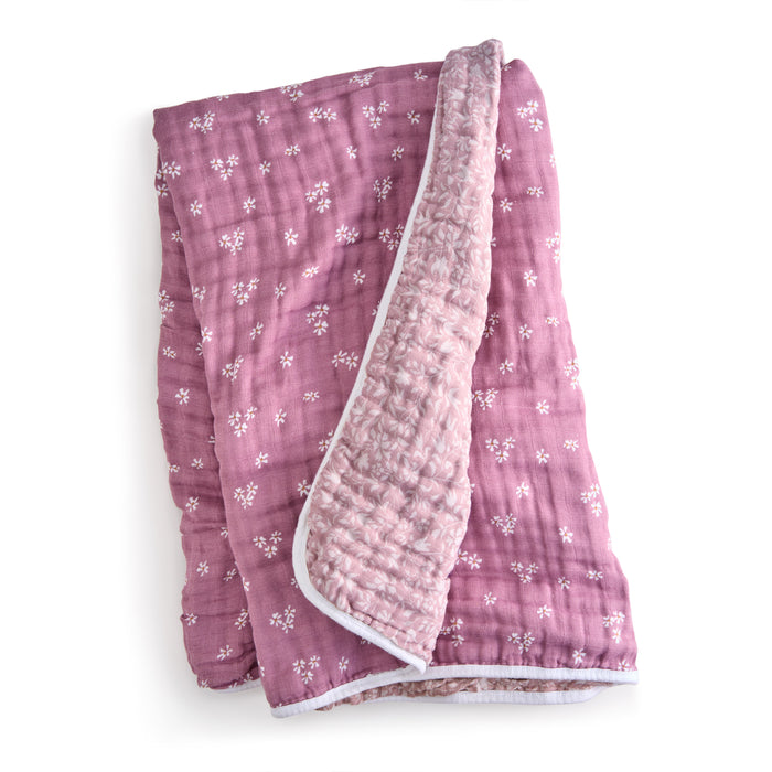 A partially folded aden + anais organic cotton muslin dream blanket, soft pink with white floral patterns, rests on a pristine background. Made from GOTS certified cotton muslin, it features a gentle textured finish inside.