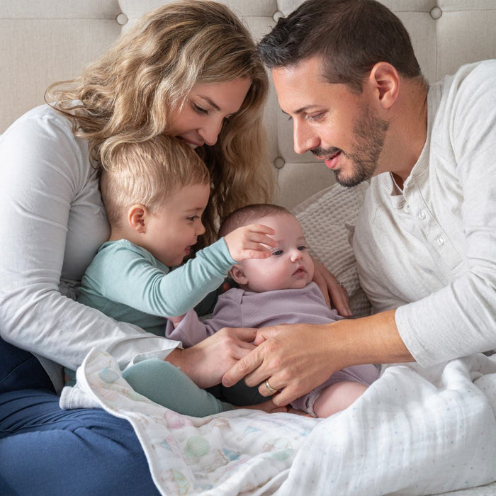 A family of four sits together on a beige couch, wrapped in an aden + anais organic cotton dream blanket. The two adults and their young children snuggle under the light-colored soft muslin cover while one child gently touches the babys head and the parents smile affectionately.