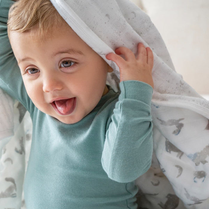 A baby with light brown hair and a playful smile sticks out their tongue while playing peekaboo, partially hidden under an aden + anais organic cotton dream blanket. The GOTS certified muslin features a subtle animal print, complementing the babys light blue outfit.