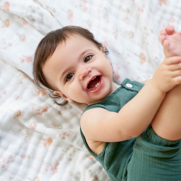 A smiling baby in a green outfit lies on an aden + anais organic cotton dream blanket, holding their foot with one hand.