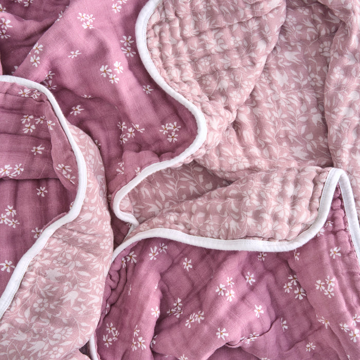 Aden + anais organic cotton muslin dream blanket is a crumpled, reversible quilt made from GOTS certified cotton. It boasts floral patterns with small white flowers on pink on one side and a subtler design on lighter pink on the other, finished with a white border.