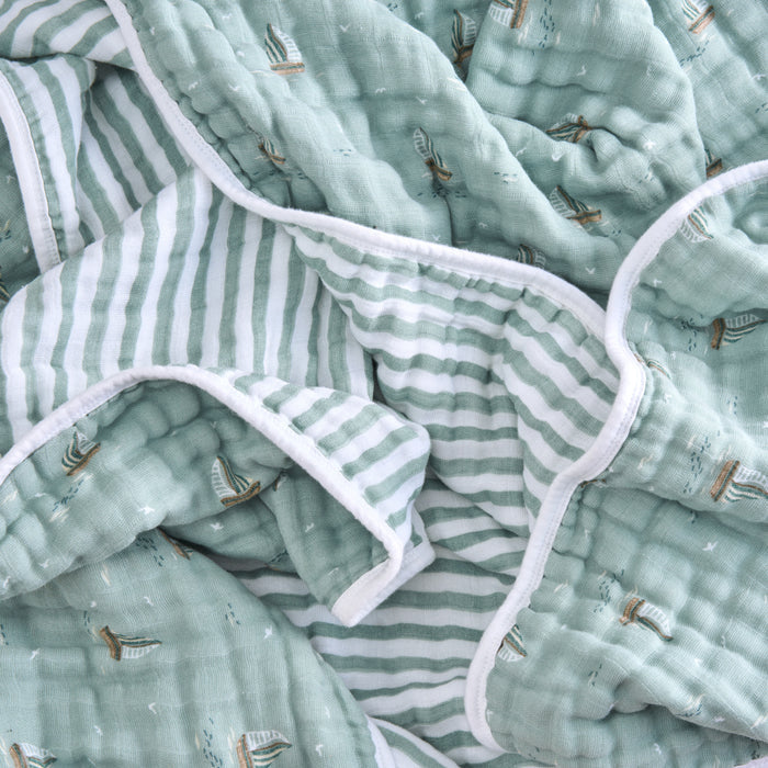 Close-up of a crumpled fabric with a nautical theme. The aden + anais organic cotton muslin dream blanket is light green with small sailboat prints and white stripes, featuring soft cotton muslin and white trim. Its GOTS certified for pure comfort.