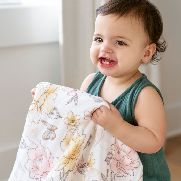 A smiling toddler with short dark hair clutches an aden + anais organic cotton dream blanket, GOTS certified and floral-patterned. The child wears a sleeveless green top while standing in a brightly lit room, wrapped in the comfort of the soft cotton muslin fabric.