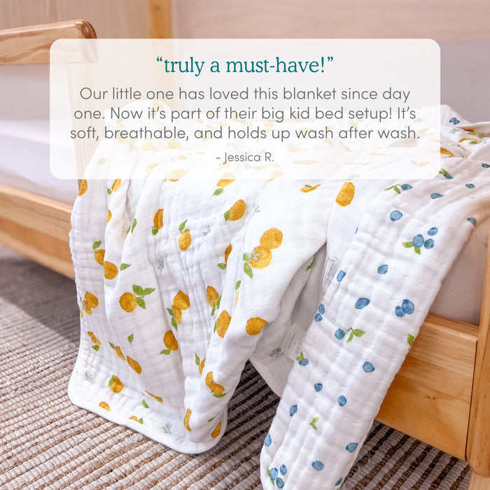 An aden + anais organic cotton muslin dream blanket with yellow lemon and blue berry prints drapes over a light wooden crib, adding charm to the bedding. A glowing review for these blankets is featured at the top of the image.