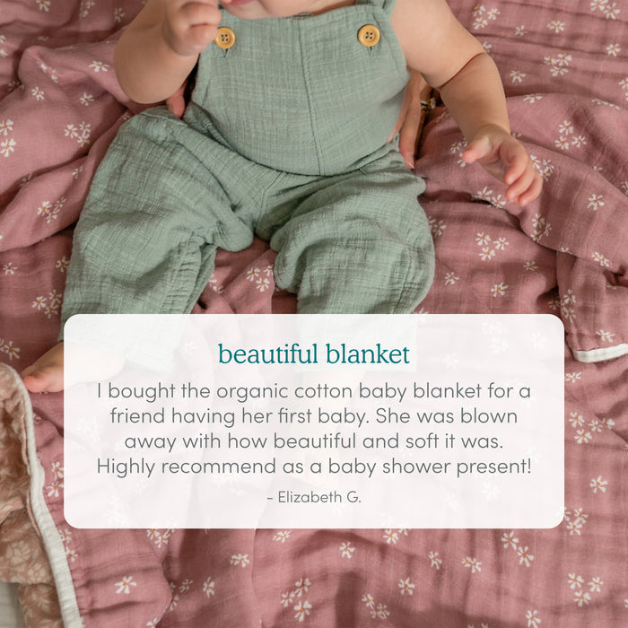 A baby in green overalls sits on an aden + anais organic cotton muslin dream blanket with a pink pattern. A review calls this blanket soft, beautiful, and a top pick for baby shower gifts.