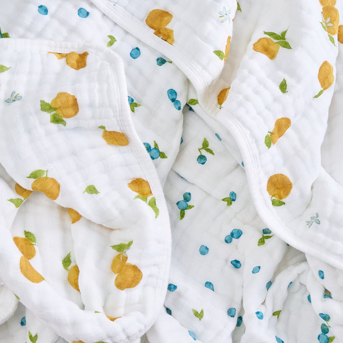 The aden + anais organic cotton muslin dream blanket is made from GOTS-certified crinkled white fabric, featuring small, colorful yellow oranges, blue berries, and green leaves. Its soft against sensitive skin and perfect for your baby.