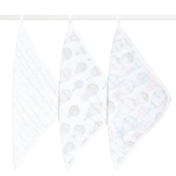 Three aden + anais organic cotton muslin oversized washcloths hang on a white rod, each boasting unique pastel patterns: one with small stars, another with hot air balloons, and the last with planets and clouds, all set against a simple white background.