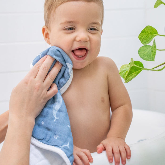 A baby in a bathtub smiles with its tongue out as someone gently wipes their face using an aden + anais organic cotton muslin oversized washcloth, which is eco-friendly, blue, and has white patterns. A green plant decorates the background against a white tiled wall.