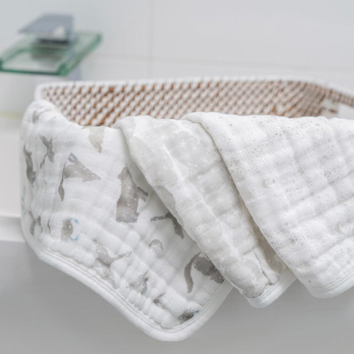 An Organic Baby Essentials Bundle by aden + anais features a soft, white organic baby blanket with a light gray animal pattern draped over a white crib. The crib has a brown woven interior, and in the blurred background sits a soap dispenser on a glass shelf.