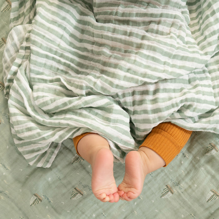 A babys feet peek from under an aden + anais organic cotton muslin swaddle blanket in green and white stripes. The baby wears orange socks, with the eco-friendly blanket resting on a light green fabric, showing that sustainable baby products can be both stylish and comforting.