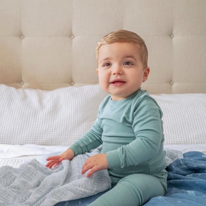 A baby in a green outfit sits on a bed with an aden + anais organic cotton muslin swaddle blanket. A beige tufted headboard and white bedding create a cozy backdrop, highlighting the sustainable swaddles.