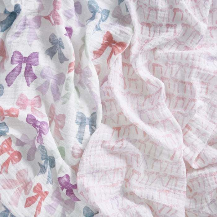Close-up of aden + anais organic cotton muslin swaddle blanket 2 pack: one with colorful bows in pink, purple, blue, and green, and one with small light pink bows. Both feature a soft, airy texture ideal for gentle snuggles.