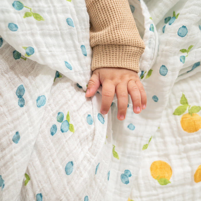 A small childs hand rests on a white blanket with blue and yellow fruit prints. The child is in a beige, waffle-patterned long-sleeve shirt, enjoying a cozy moment atop an aden + anais organic cotton muslin swaddle blanket from the 2 pack set.