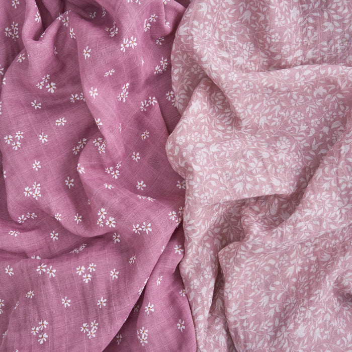 Close-up of overlapping pieces of aden + anais organic cotton muslin swaddle blankets: the left is deep pink with small white florals, and the right is lighter pink with denser white leaves, creating a textured contrast.